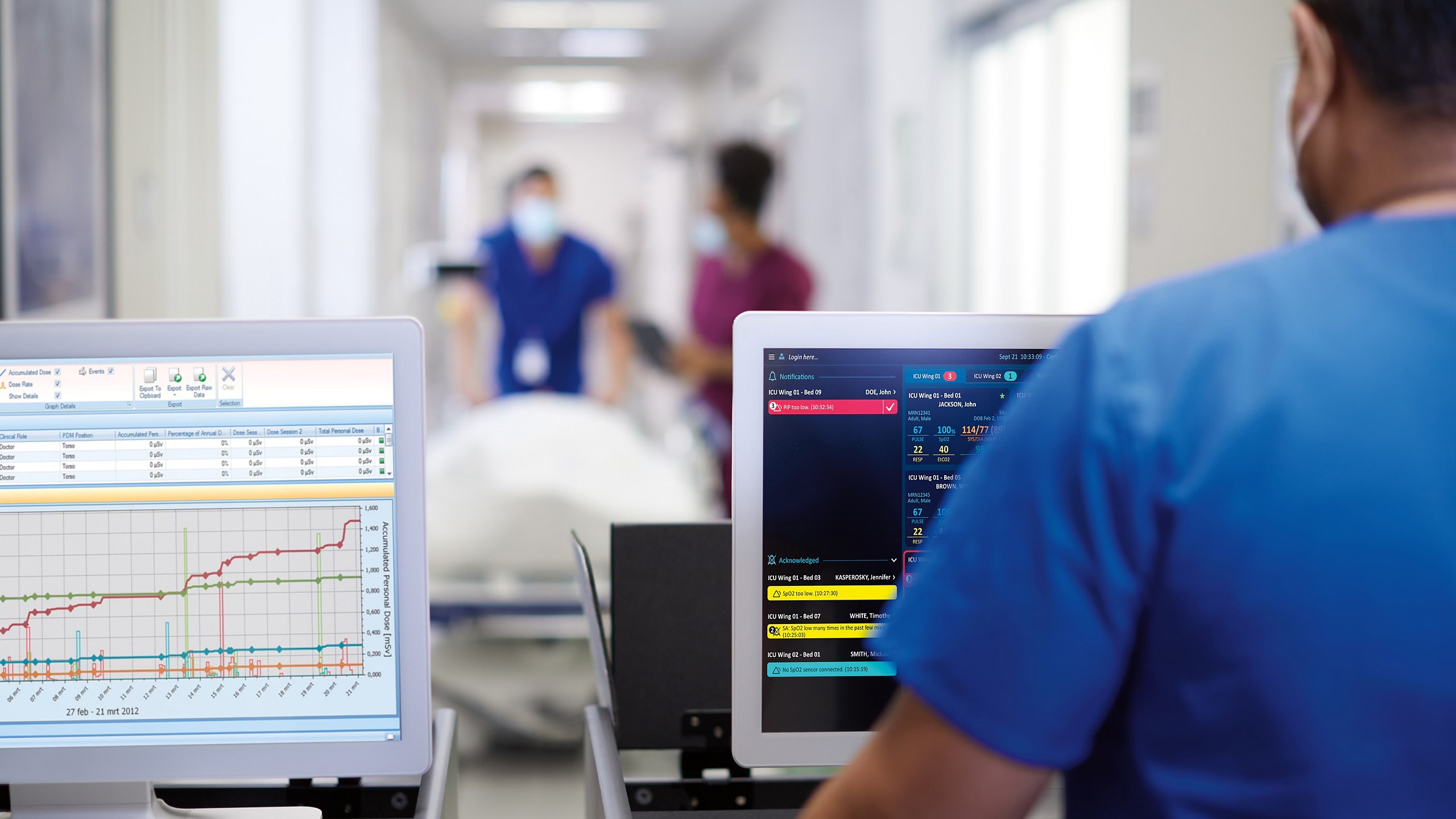 A view over the shoulder of a male medical worker, wearing scrubs and a mask, looking at two computer screens displaying critical patient data, including vital signs and graph trends, in a busy hospital hallway.