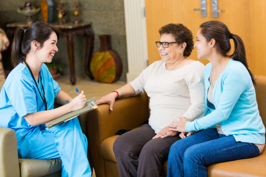 A smiling nurse in blue scrubs is sitting on a sofa and writing on a clipboard while consulting with an older adult woman and a younger female relative, all smiling and engaged in a friendly discussion about care options.