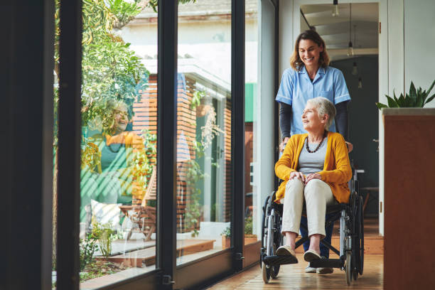 A smiling nurse in blue uniform is gently pushing a happy senior woman in a wheelchair down a bright, sunlit hallway near a large window, suggesting assisted living or home care.