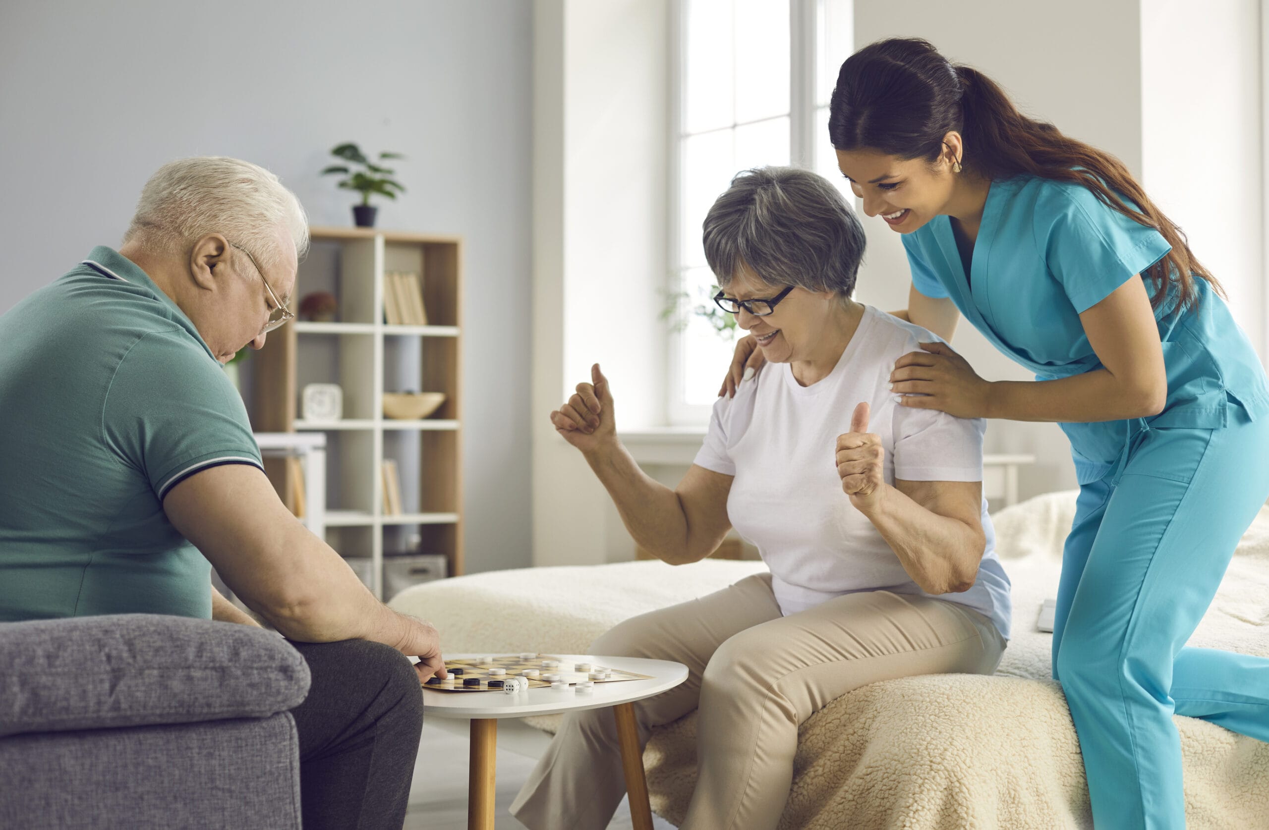 A female nurse is cheering on a delighted senior woman who gives a thumbs-up while sitting with an elderly man at a small table, playing a board game in a brightly lit residential care setting.